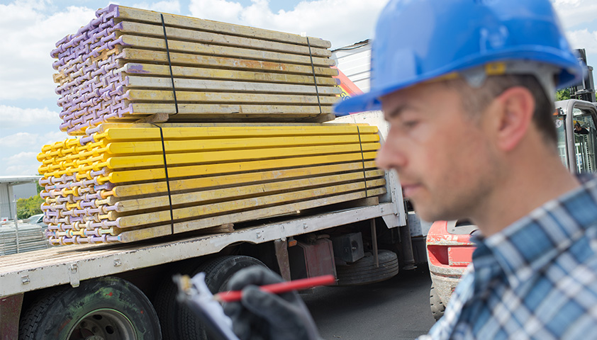 MORAVIA Akademie Aktuell: So sichern Sie Ihre Ladung Bauarbeiter mit blauem Schutzhelm inspiziert auf einer Baustelle eine LKW-Ladung ordentlich gestapelter gelber und violetter Gerüstbretter.