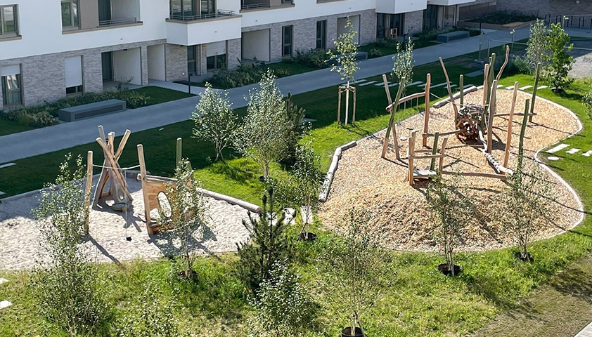 MORAVIA Akademie Aktuell: Unsere neuen Seminare zur Spielplatzkontrolle Moderner Spielplatz im Freien mit hölzernen Klettergerüsten, umgeben von jungen Bäumen und viel Grün in einem Wohngebiet.