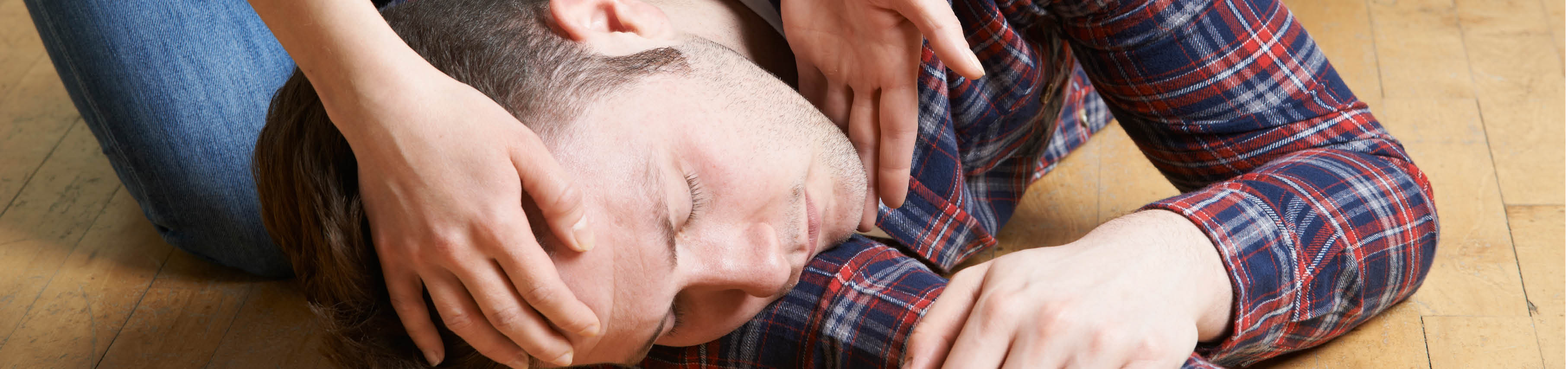 Tag der Ersten Hilfe 2024: Ein Tag zum Retten und Helfen Eine Person liegt auf der Seite auf dem Boden und scheint bewusstlos oder unwohl zu sein, während eine andere Person Hilfe leistet, indem sie ihren Kopf stützt und ihren Zustand überwacht.