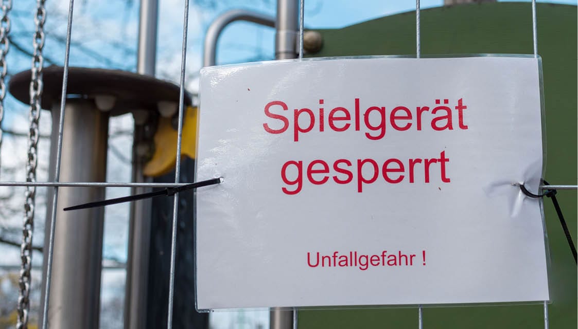 Schild "Spielgeräte gesperrt Unfallgefahr" an Drahtzaun mit Spielplatz im Hintergrund.