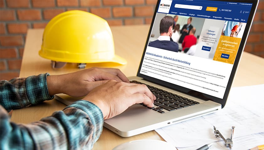 MORAVIA Akademie Aktuell: Entdecken Sie unsere neue Website - jetzt Angebote sichern mit exklusivem Rabatt Person, die einen Laptop mit der Moravia Webseite auf dem Bildschirm, einem Schutzhelm auf dem Tisch und Sicherheit am Arbeitsplatz verwendet.
