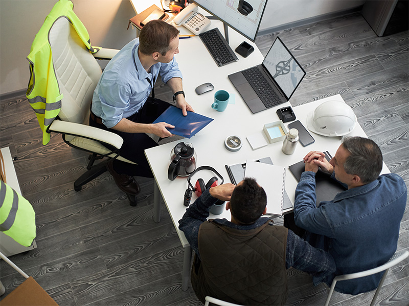 Drei Personen besprechen Bauunterlagen an einem Schreibtisch mit Laptop, Bauhelm und Warnweste im Büro