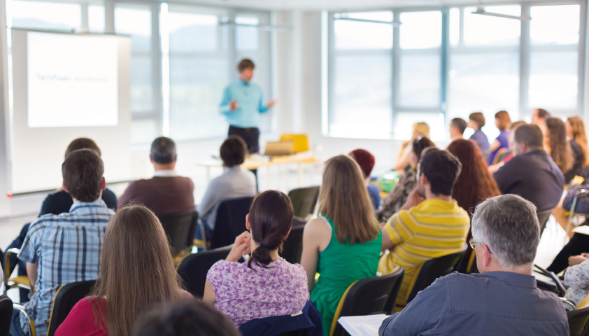 MORAVIA Akademie Aktuell: Neue Seminare, neue Broschüre Gruppe von Personen, die an einer Präsentation in einem hellen, modernen Konferenzraum teilnehmen.