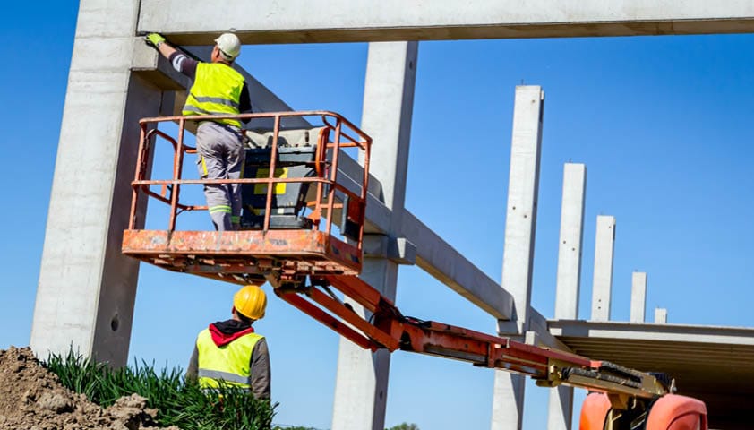 Ein Arbeiter steht auf einem Teleskopstapler auf einer Baustelle und ein weiterer Arbeiter schaut zu ihm nach oben